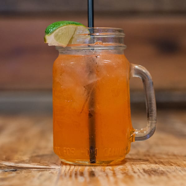 A mason jar mug filled with an orange drink, ice, a black straw, and a lime wedge.
