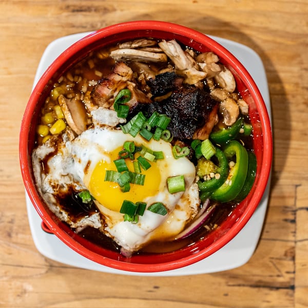Overhead view of a red bowl of ramen with a fried egg, meat, corn, and jalapeños.