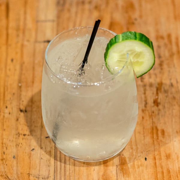 A refreshing cocktail with ice, a black straw, and a cucumber slice on a wooden table.